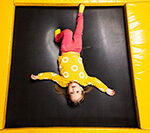 Girl in yellow sweater laying down and looking up at camera while laying on a trampoline at indoor playground near homes for sale in Howell NJ