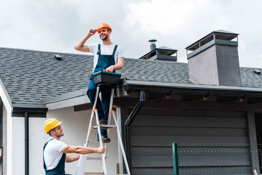 Chimney service Hazlet technicians in hard hats using a ladder to reach a roof with two capped chimneys